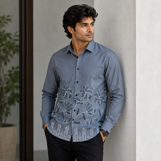 A man wearing a slate blue long-sleeve button-up shirt with white floral embroidery across the lower half, paired with black trousers, standing against a light gray wall.